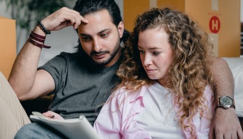 A couple is sitting next to boxes and looking at a notebook.